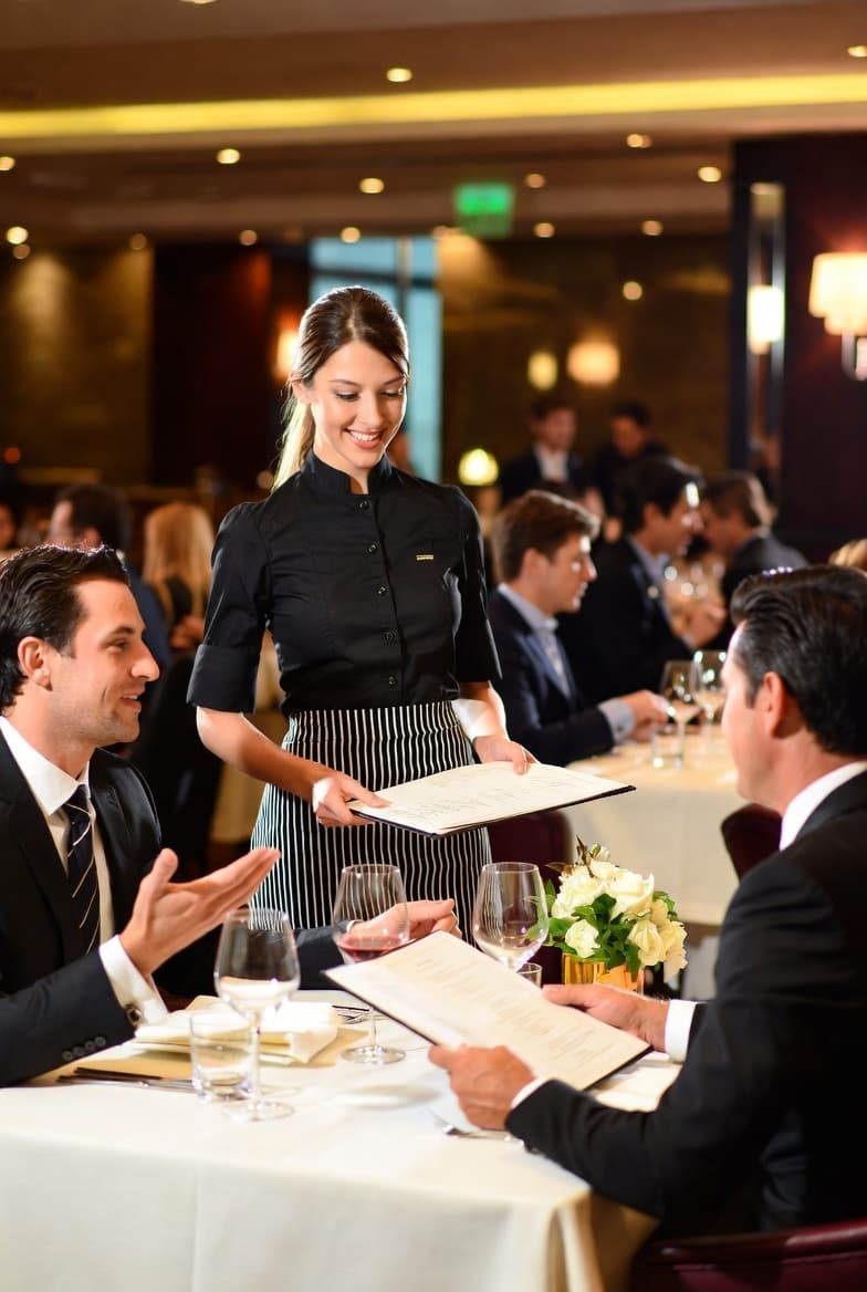 Server presenting a wine list to seated guests in an upscale dining room with a wine wall
